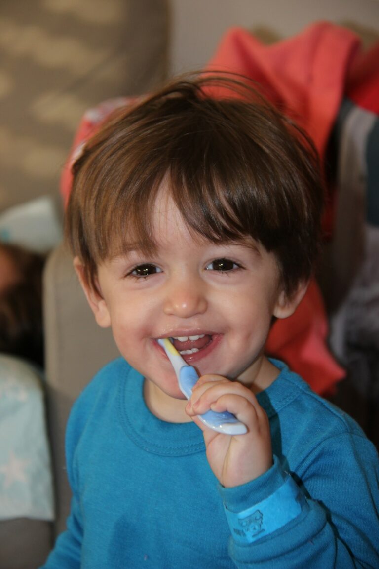 A young child stands at a bathroom sink, smiling and brushing teeth with a parent supervising nearby.