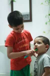 Parent helps a young child brush primary teeth at a bathroom sink.